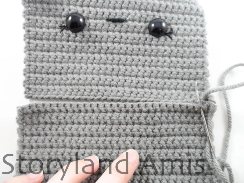 Two gray crocheted rectangular panels are being sewn together with a silver tapestry needle to create an amigurumi robot head.