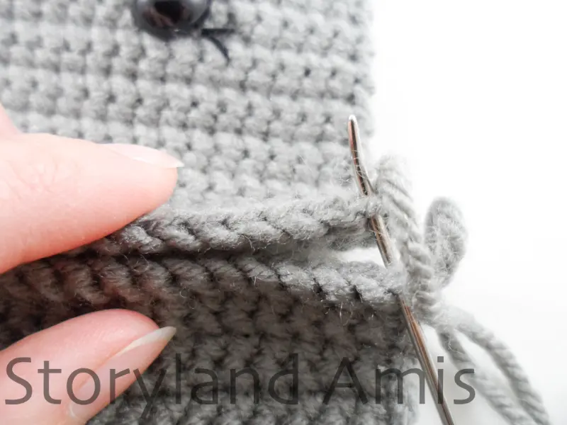 A close-up of two gray crocheted rectangles being sewn together with a silver tapestry needle.