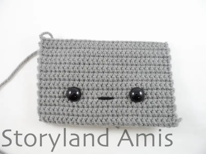 A gray crocheted rectangle lies on a white background. In the center of the rectangle are two black plastic safety eyes. Between the two eyes is a black straight stitch.