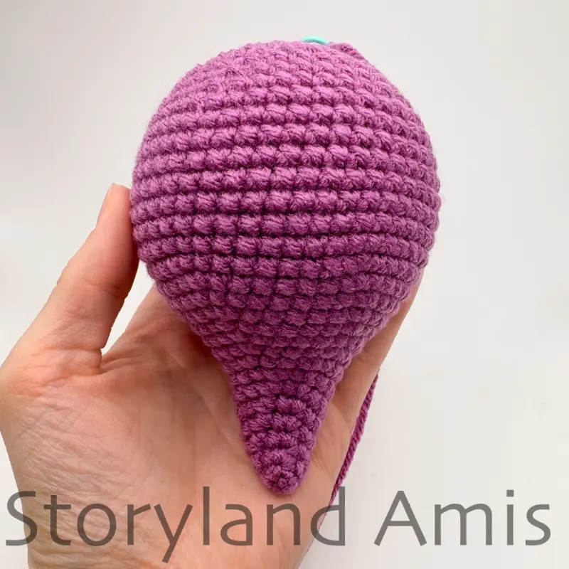 A hand holds up a partially finished amigurumi stegosaurus dinosaur body, showing it from above.