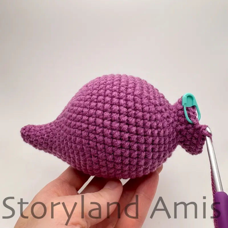 A hand holds up a partially finished amigurumi stegosaurus body and head. A purple crochet hook is inserted into a stitch of the head.