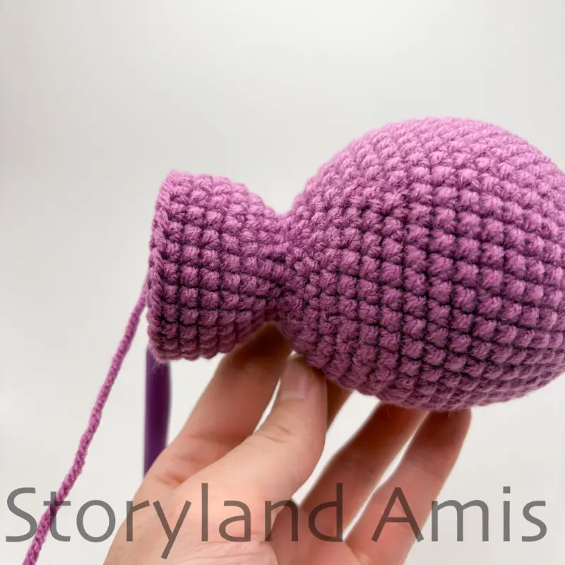 A hand holds up a partially finished purple amigurumi stegosaurus.