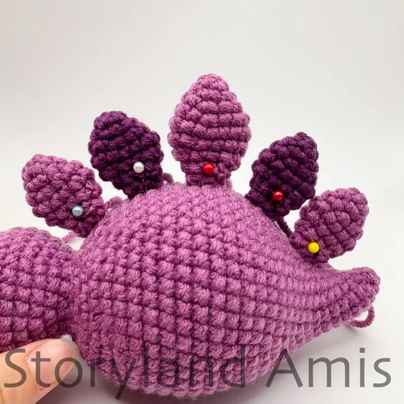 A hand holds up a partially finished amigurumi stegosaurus. The plates on the dinosaur's back are pinned into place with straight pins.