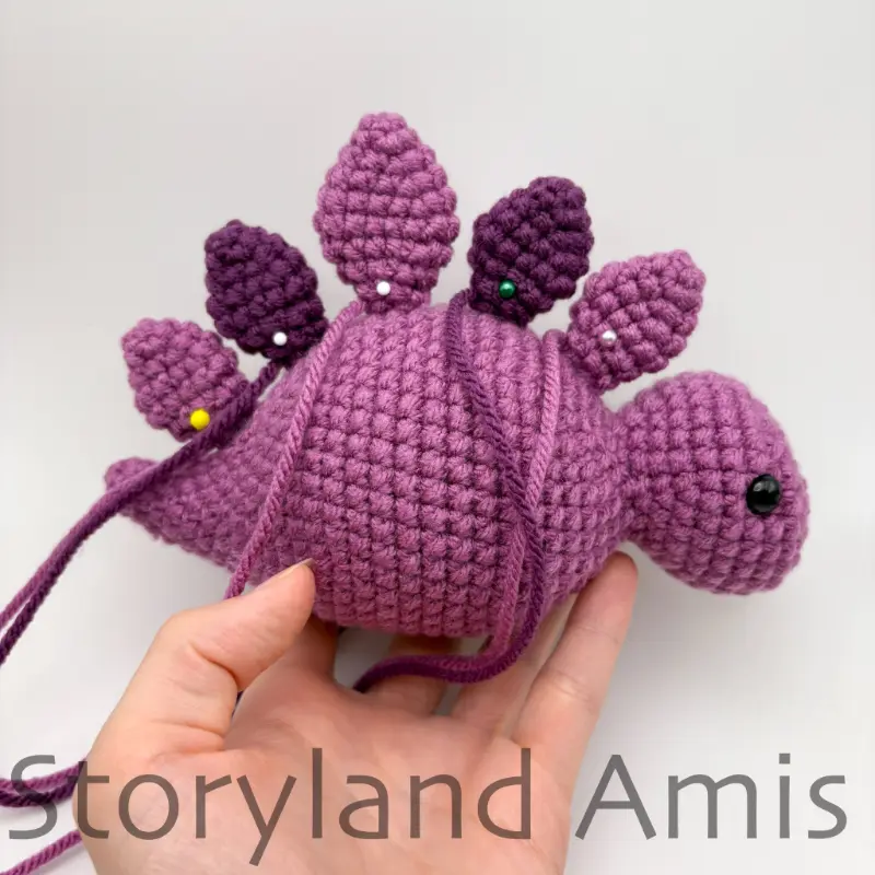 A hand holds up a partially finished amigurumi stegosaurus. The plates on the dinosaur's back are pinned into place with straight pins.