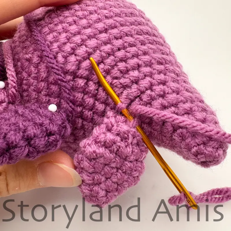 A close-up of a purple plate being sewn to the back of an amigurumi stegosaurus with a gold tapestry needle.