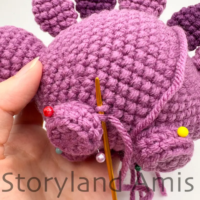 A close-up of a crocheted leg being sewn to the body of an amigurumi stegosaurus using a tapestry needle.
