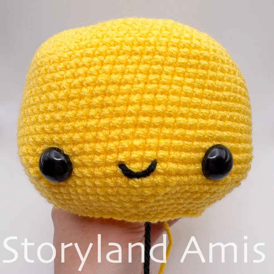 A hand holds up a partially finished bee head with two black plastic safety eyes and a mouth. 