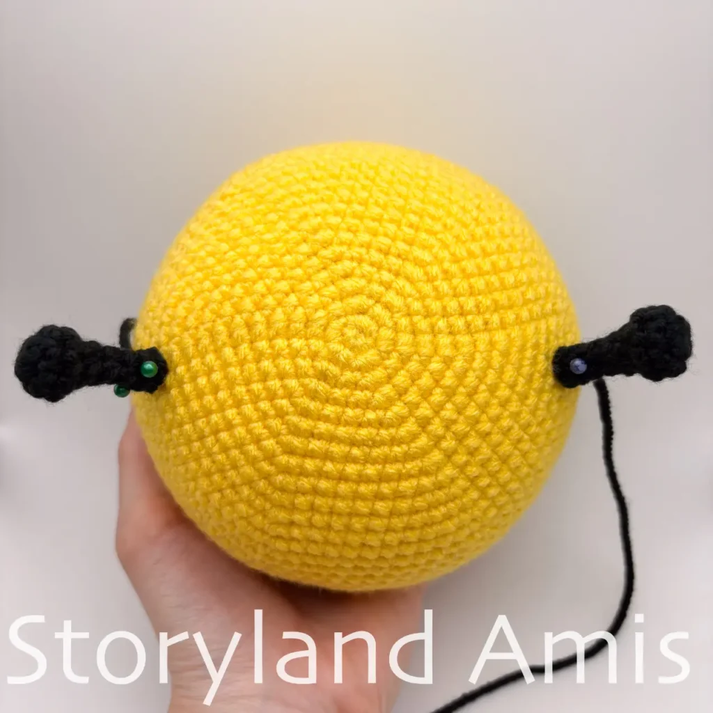 An amigurumi bee head is shown from the top. Two crocheted black antennas are placed on either side of the head and pinned in place with straight pins.