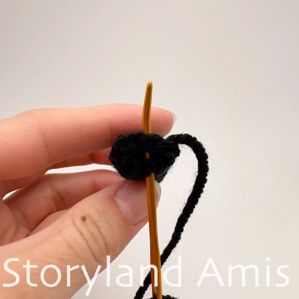 A hand holds a black crocheted bee arm. A tapestry needle is inserted into the stitches of the last round of the arm. 
