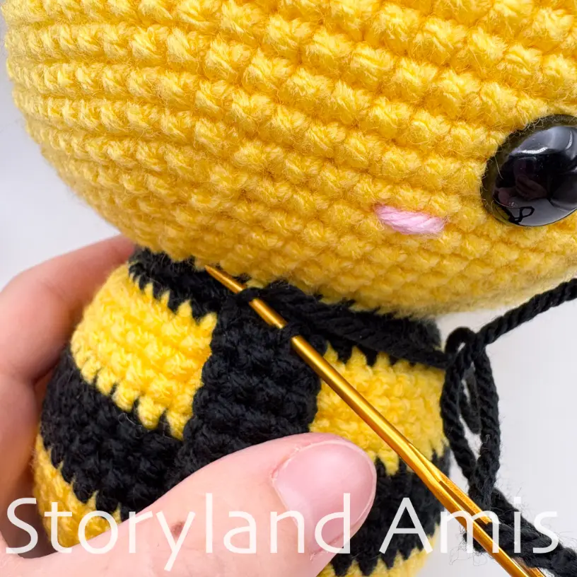 A close-up of a crocheted arm being sewn to the body of an amigurumi bee with a tapestry needle. 