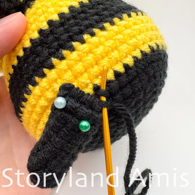 A close-up of a crocheted leg being sewn to the body of an amigurumi bee using a tapestry needle.