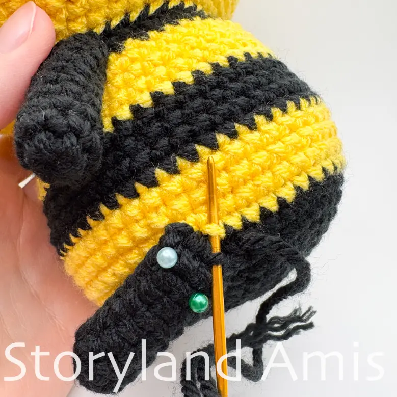 A close-up of a crocheted leg being sewn to the body of an amigurumi bee using a tapestry needle.