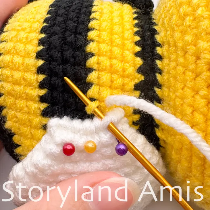 A close-up shows a hand using a tapestry needle to sew a crocheted wing to the back of the body of an amigurumi bee.