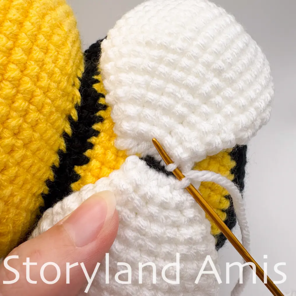 A close-up shows a hand using a tapestry needle to sew a crocheted wing to the back of an amigurumi bee's body.