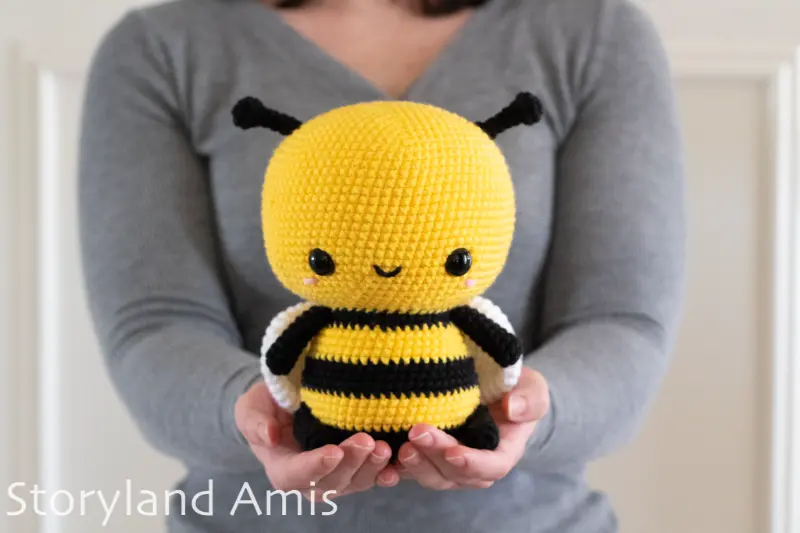 A girl wearing a gray sweater holds a large crocheted amigurumi bee.