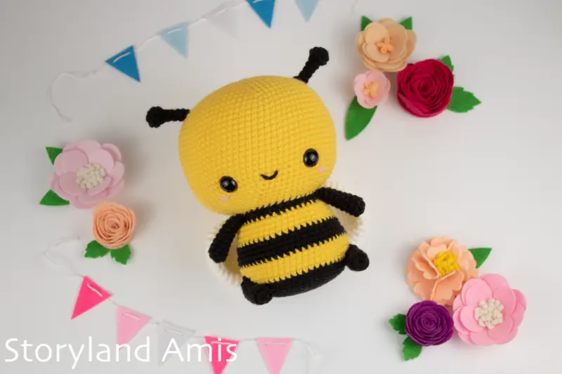 An amigurumi bumble bee lies on a white table surrounded by felt flowers and colorful pendants. 
