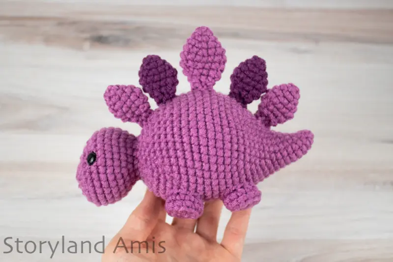 A hand holds a purple crocheted stegosaurus toy against a wooden background.