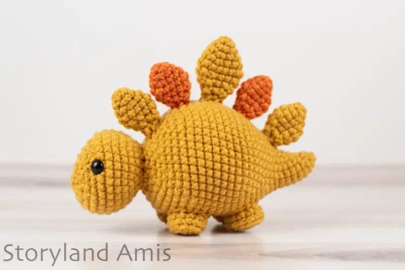 An orange and yellow crocheted stegosaurus toy sits on a wooden table.