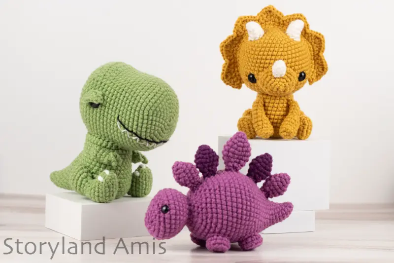 A crocheted t-rex, stegosaurus, and triceratops sit on a wooden table in front of a white background.