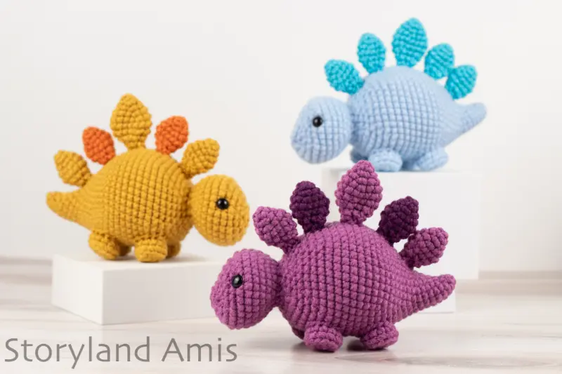 Three amigurumi stegosaurus dinosaurs stand on a wooden table.