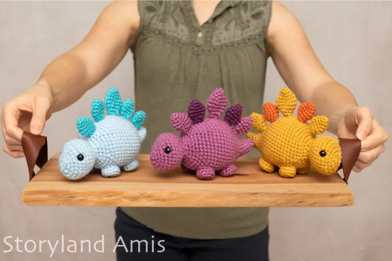 A girl holds a wooden tray with three crocheted stegosaurus amigurumi toys on it.