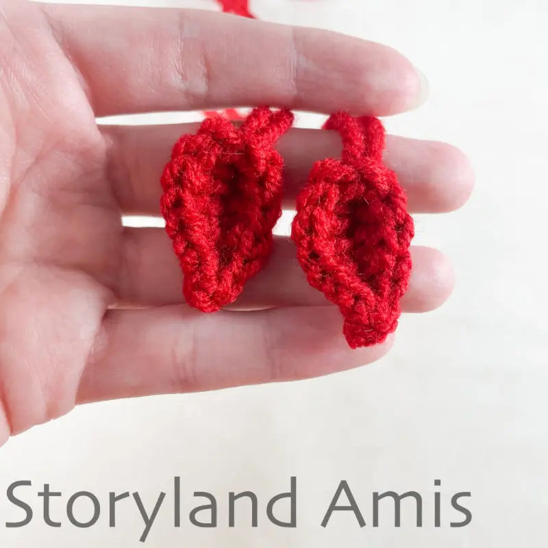 A hand holds two red crocheted pieces to be used as hat ties on a crocheted hat.