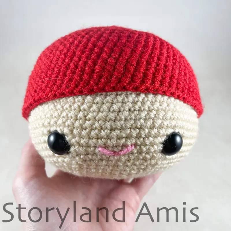 A hand holds an amigurumi head wearing a red crocheted hat.