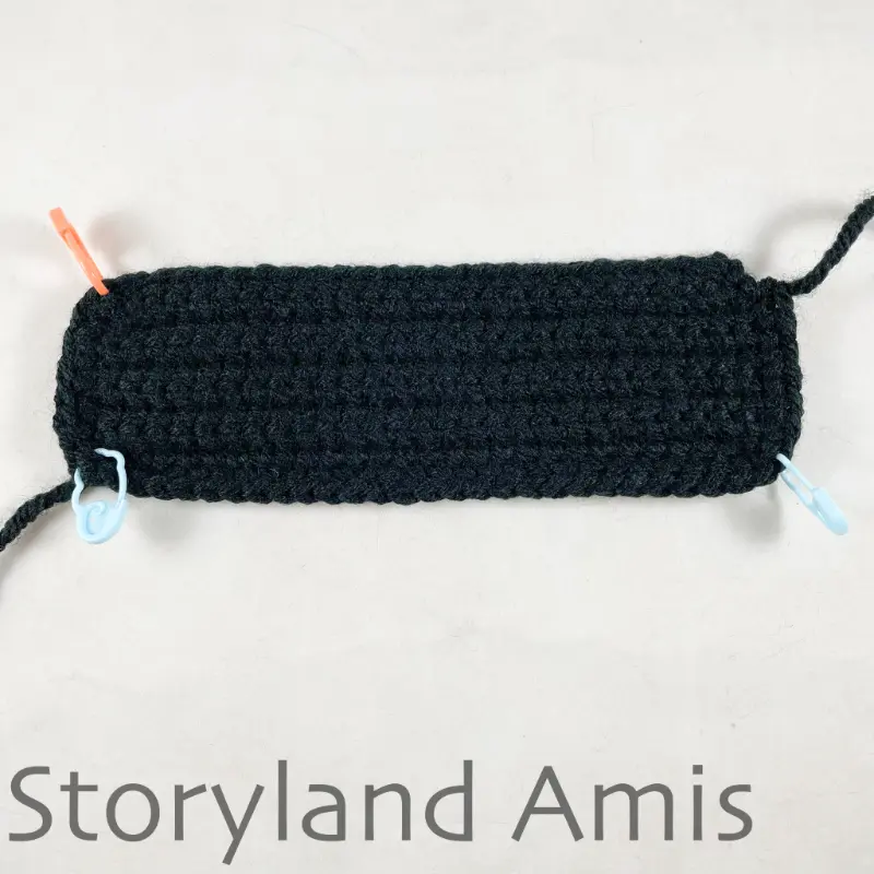 A black crocheted rectangle lays on a white table. The rectangle has a stitch marker on three of the corners.