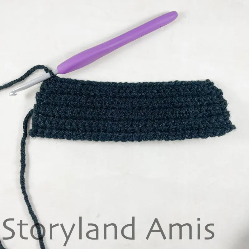 A partially finished black crocheted vest for an amigurumi pirate doll lies on a white table with a crochet hook inserted in the corner stitch.