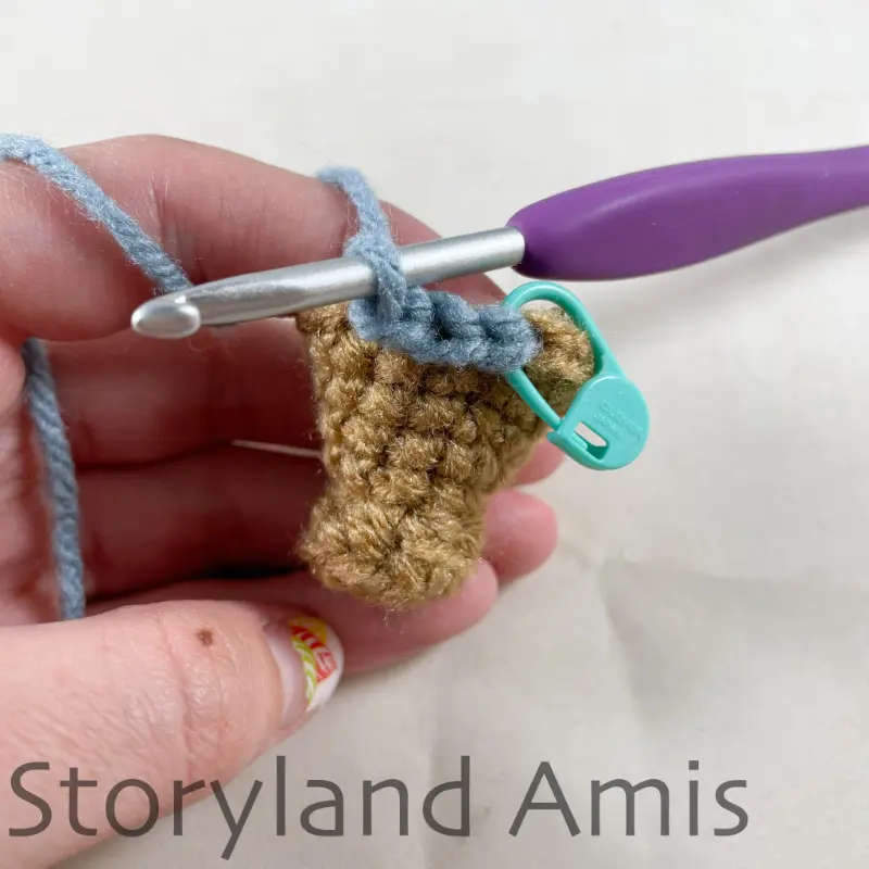 A partially finished crocheted pirate leg is held steady by a hand. A purple crochet hook is inserted in one of the stitches of the leg.
