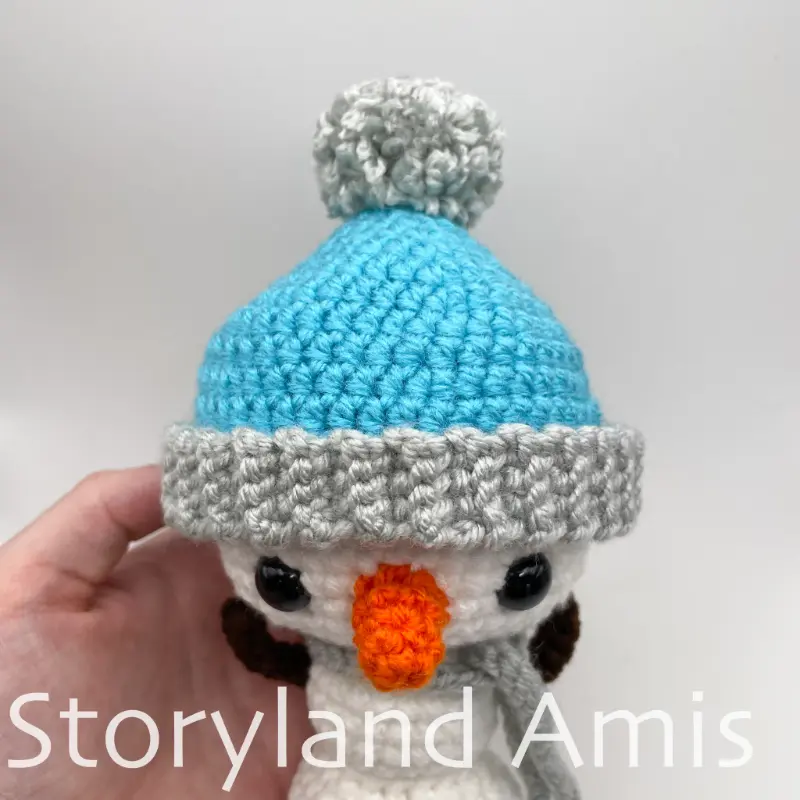 A close-up of an amigurumi snowman's head wearing a blue and gray stocking cap.