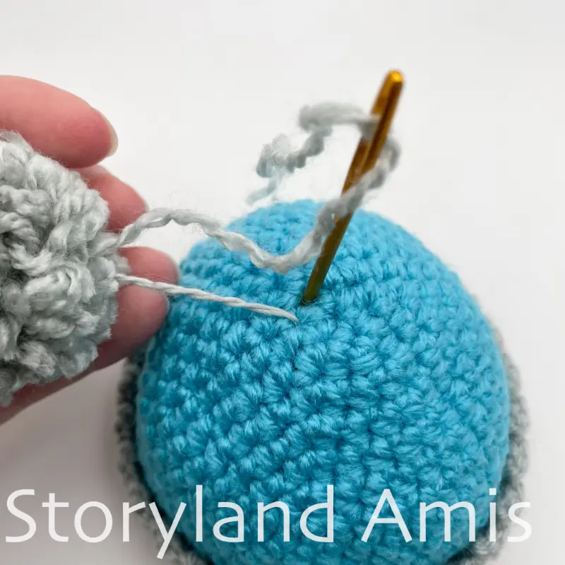 A tapestry needle is shown sewing a pom pom to the top of a blue stocking cap. A hand holds the pom pom steady.