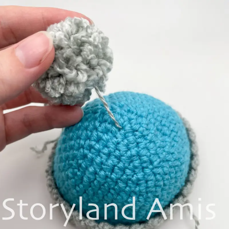 A hand shows how to add a pom pom to the top of a small stocking cap for an amigurumi snowman.