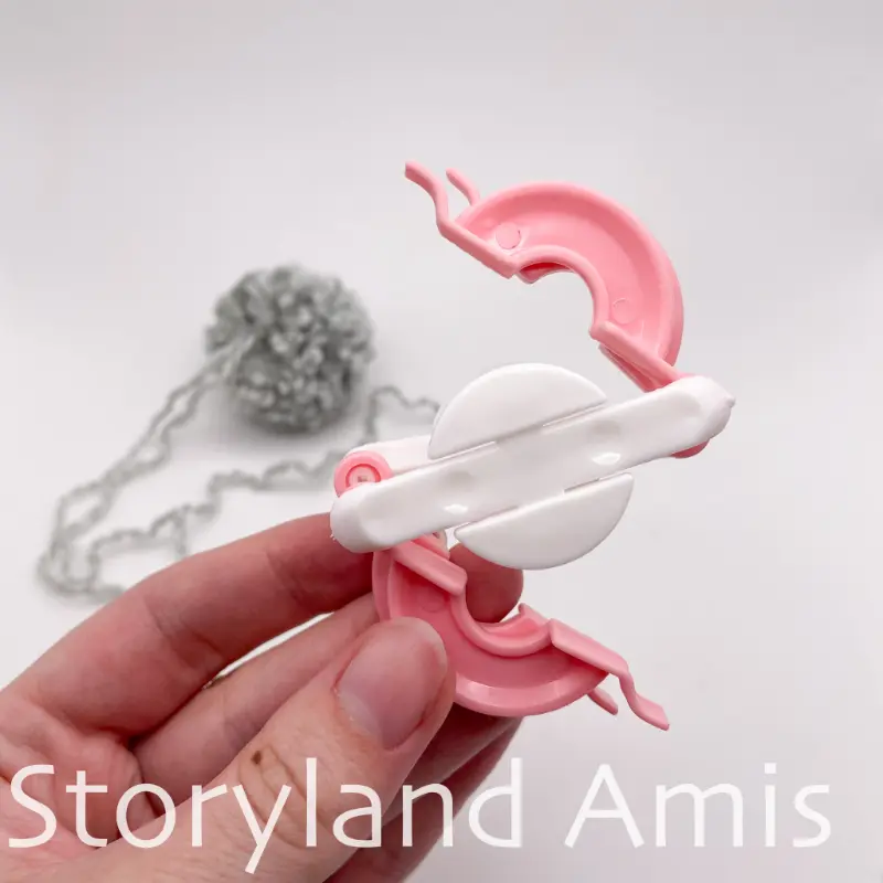 A hand holds up a pink and white pom pom maker. A gray pom pom lays on the table behind it.
