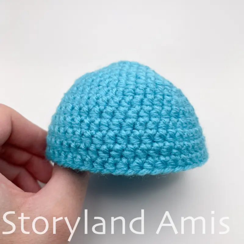 A hand holds a partially finished blue stocking cap for an amigurumi snowman.