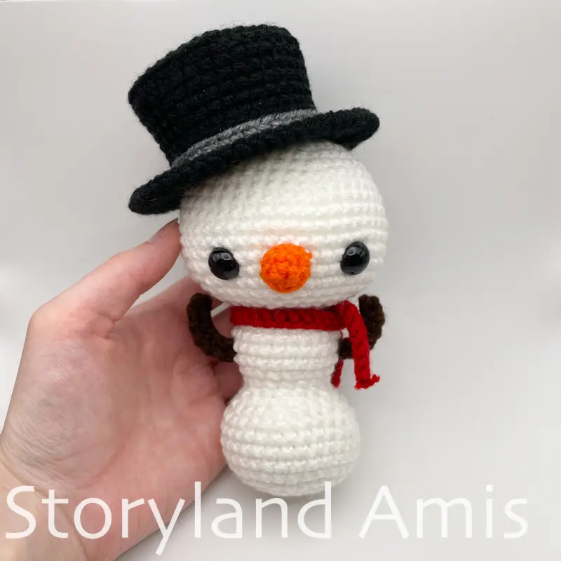 A hand holds an amigurumi snowman wearing a red scarf and a black top hat.