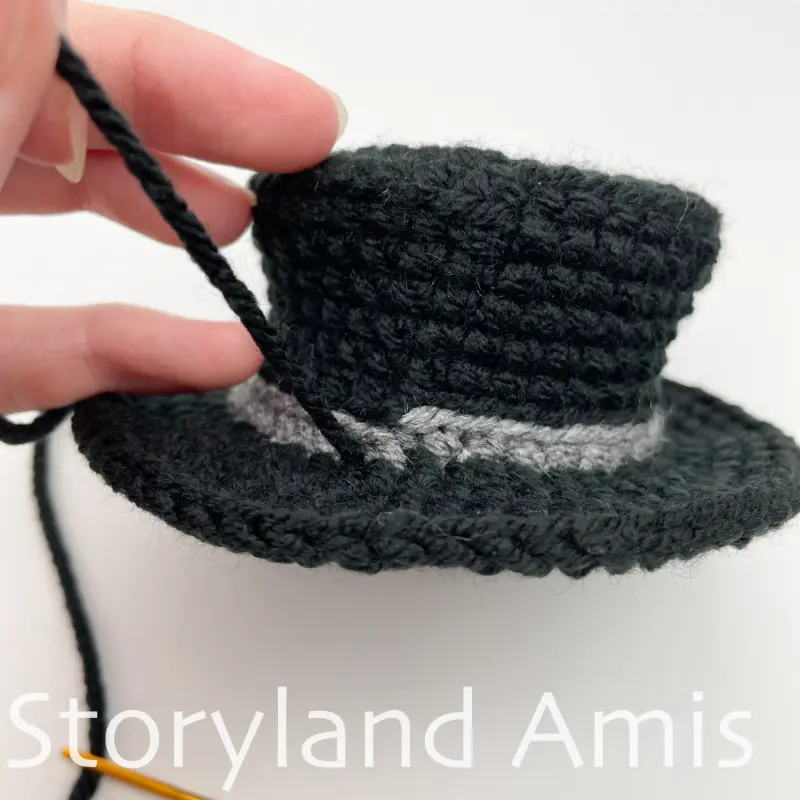 A crocheted top hat with a black yarn tail coming out of the brim.