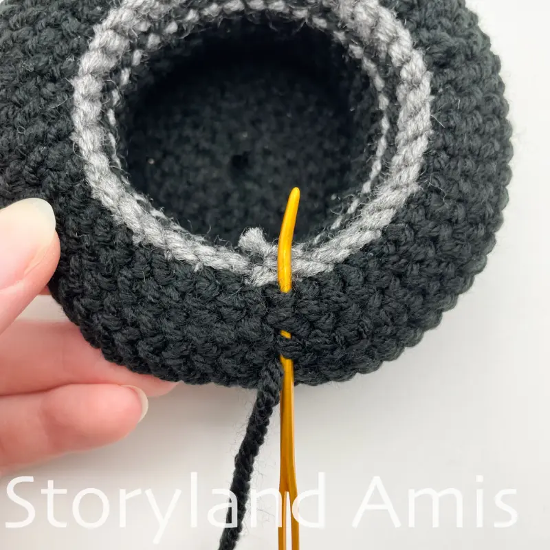 A close-up shows a tapestry needle weaving a yarn tail to the inside edge of a crocheted top hat.