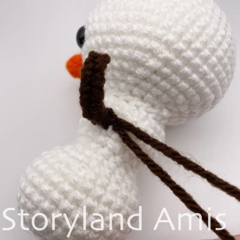 A close-up of the side of an amigurumi snowman showing the arm with two yarn tails coming out of it.