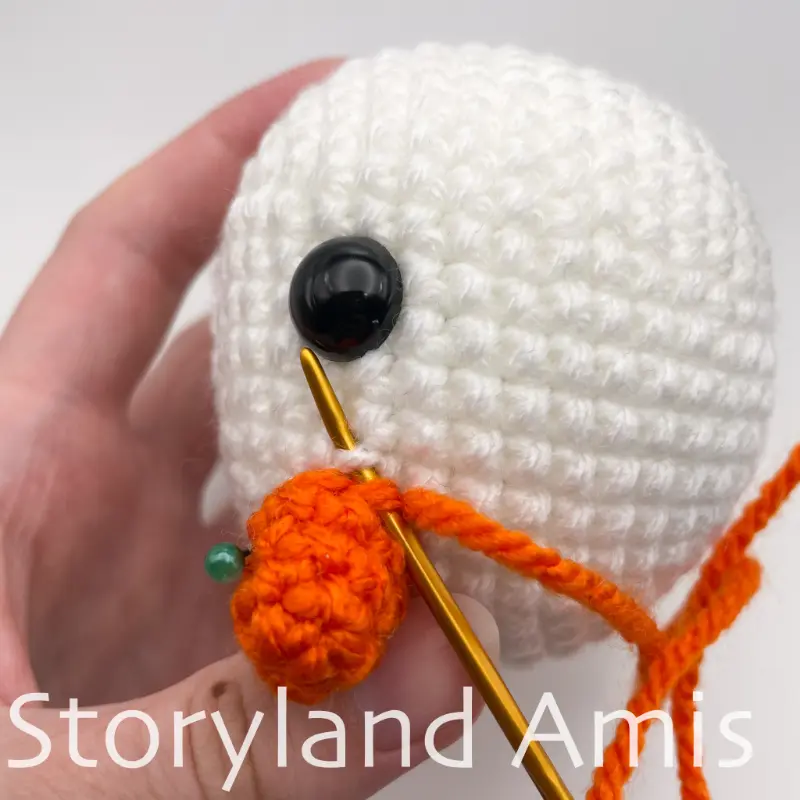 A close-up shows a crocheted carrot nose being sewn to the head of an amigurumi snowman using a gold tapestry needle.