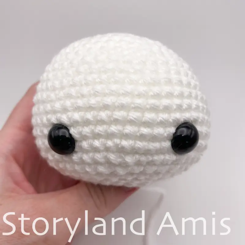 A hand holds a white, crocheted snowman head.
