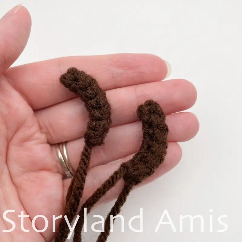 A hand holds two brown crocheted snowman arms.