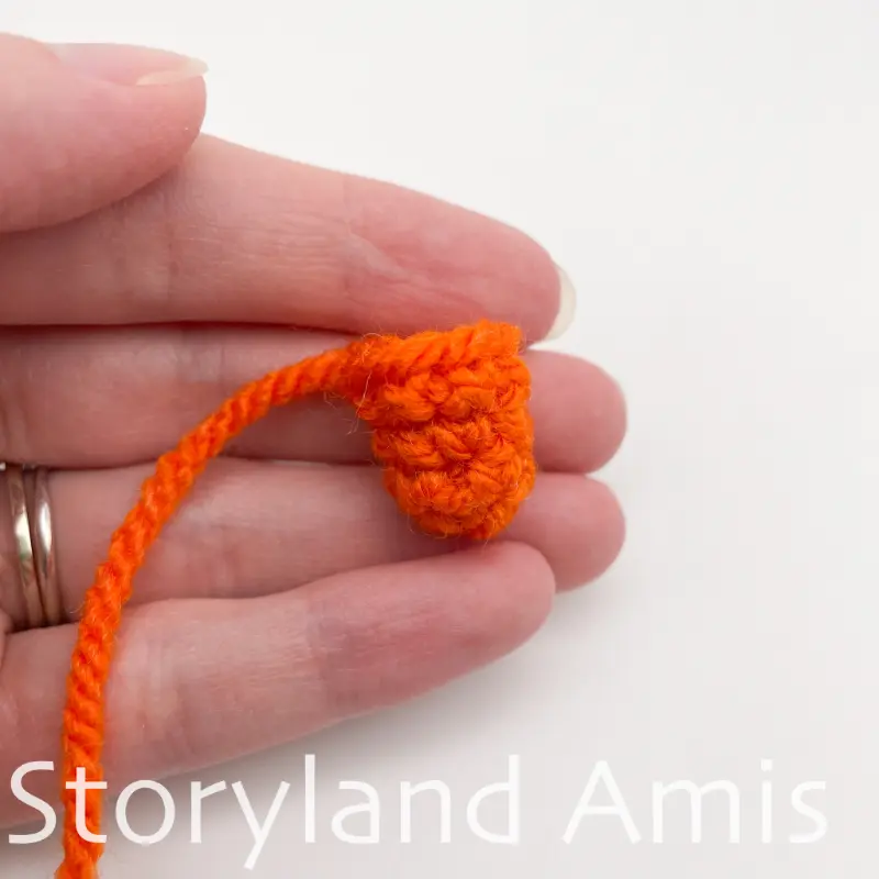A hand holds an orange crocheted carrot nose for an amigurumi snowman.