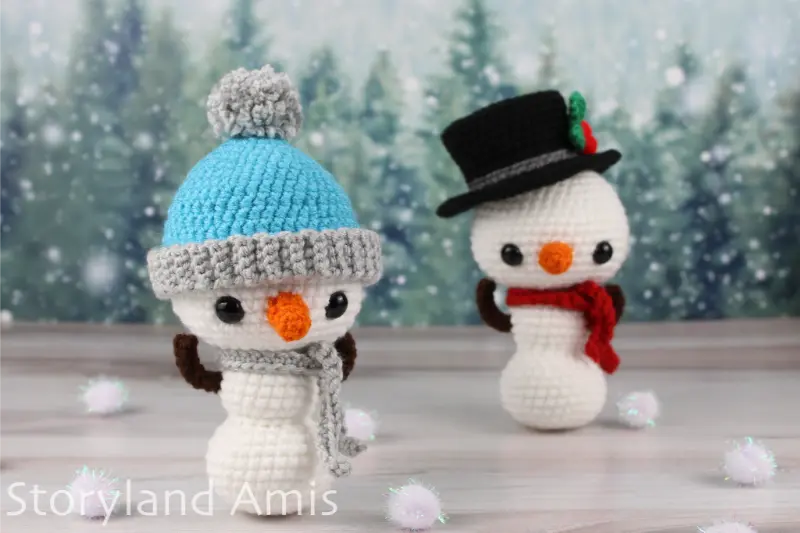 Two amigurumi snowmen stand on a wooden table with pom poms surrounding them in front of a snowy tree background.