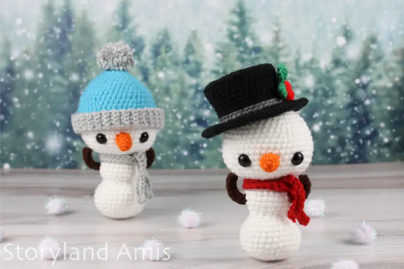 Two amigurumi snowmen stand on a wooden table with pom poms surrounding them in front of a snowy tree background.