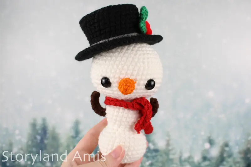A hand holds up an amigurumi snowman in front of a snowy tree background.