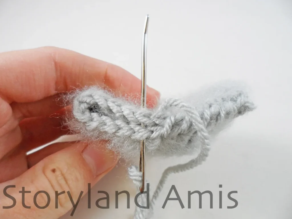 A hand uses a tapestry needle to demonstrate how to sew the open ends closed on a crocheted koala ear to create a flat edge for sewing.