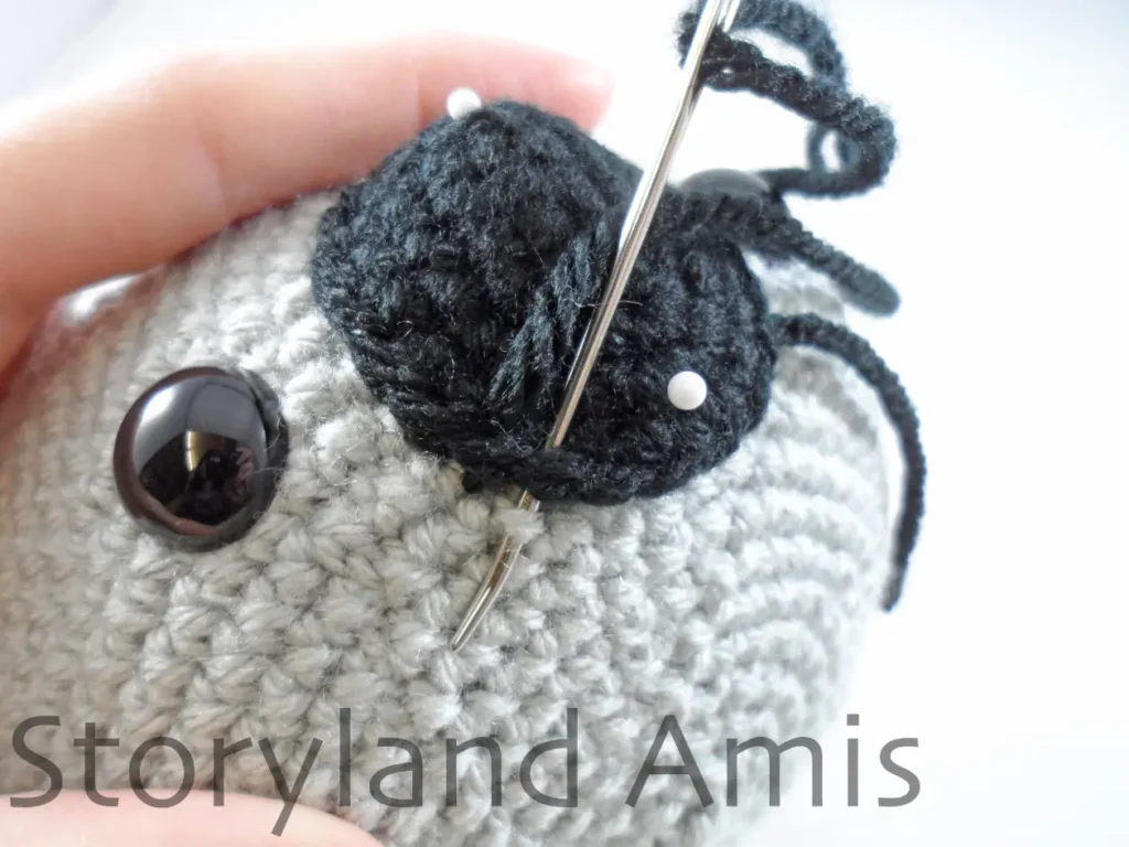 A close-up of a black crocheted nose being sewn to an amigurumi koala's head using a tapestry needle.