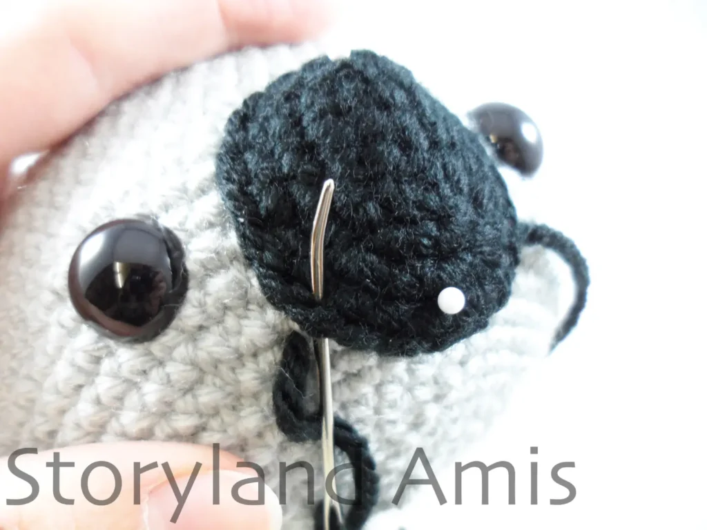 A close-up of a black crocheted nose being sewn to an amigurumi koala's head using a tapestry needle.