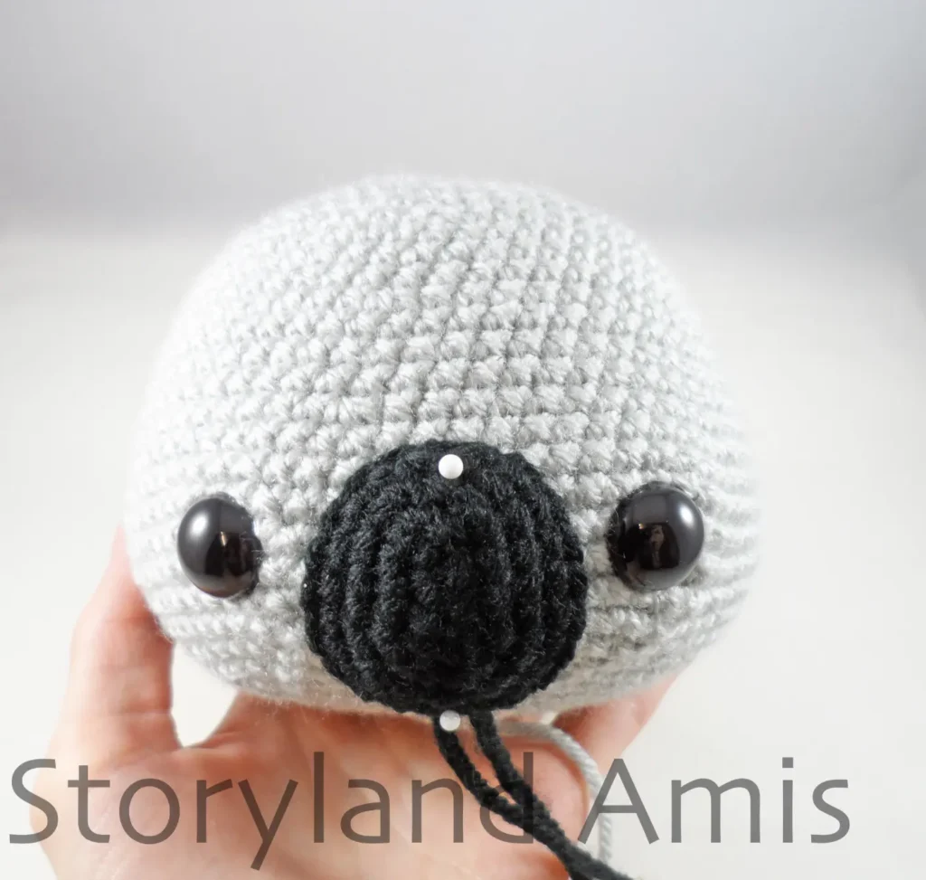 A hand holds a partially finished amigurumi koala head with a black crocheted nose pinned between its eyes.